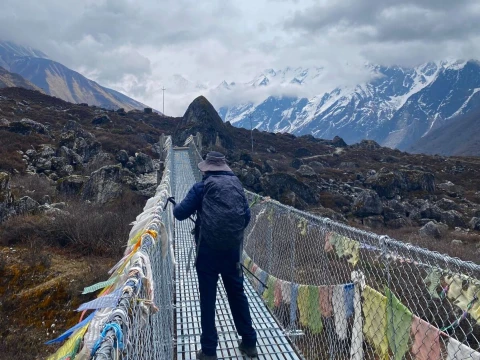 Trekker crossing a suspension bridge in Langtang Valley with cloudy skies and snow-covered Himalayan mountains.