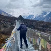 Trekker crossing a suspension bridge in Langtang Valley with cloudy skies and snow-covered Himalayan mountains.