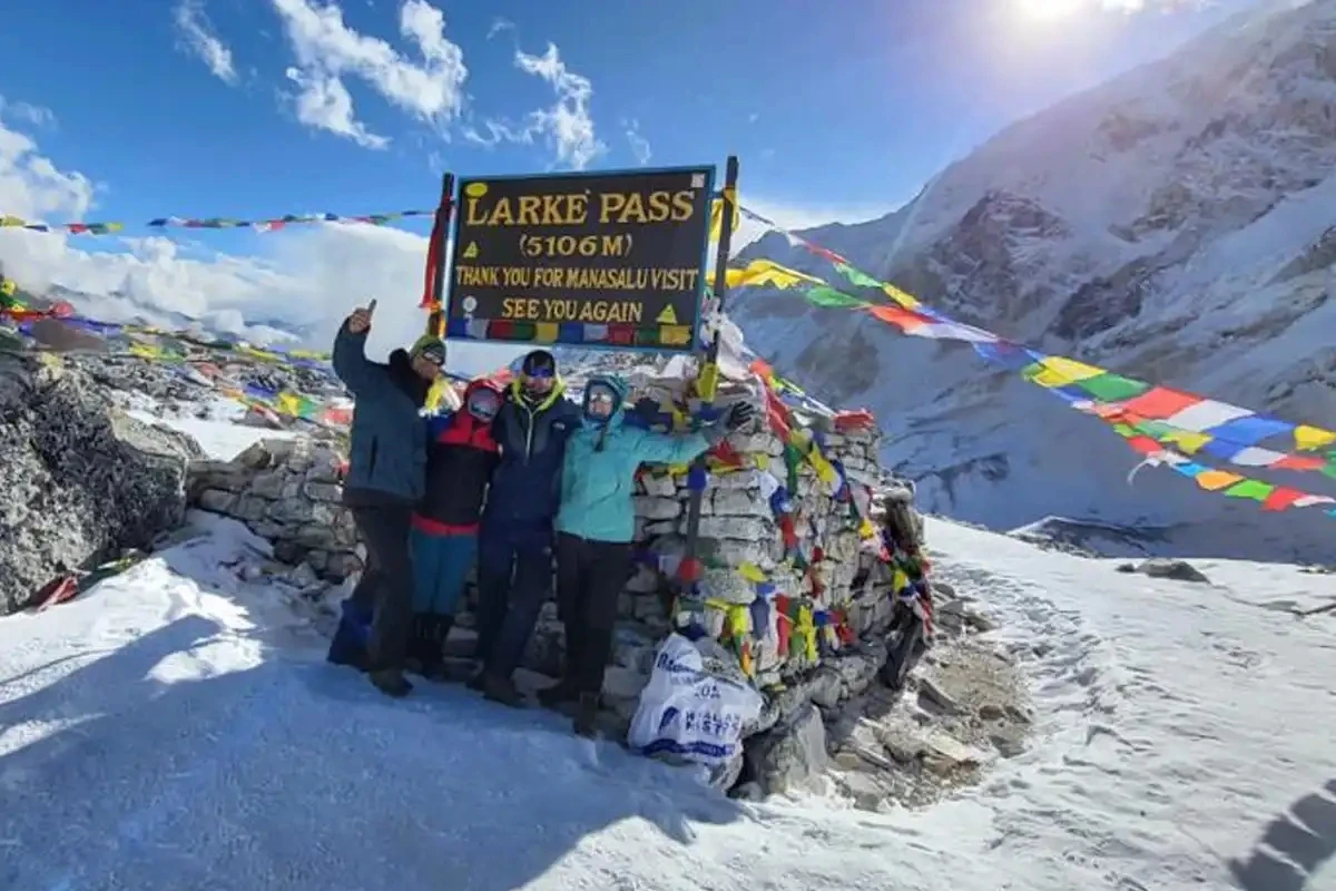 Group of trekkers posing at the Larke Pass (5106m) sign on the Manaslu Circuit Trek surrounded by snow-covered mountains and colorful prayer flags.